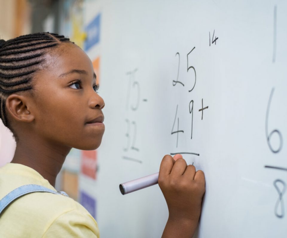 Portrait of african girl writing solution of sums on white board at school. Black schoolgirl solving addition sum on white board with marker pen. School child thinking while doing mathematics problem.
